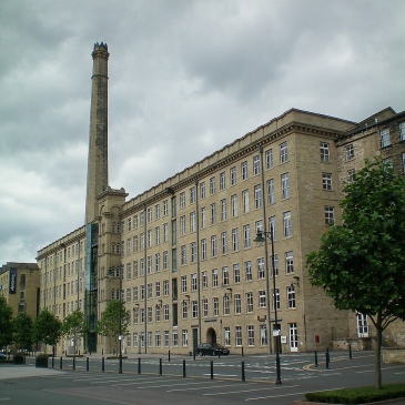 A large multi-storey mill building, each floor has many windows. A large chimney is in the centre